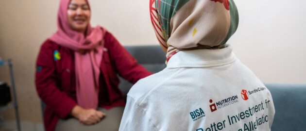 Two woman are conversing. One woman is facing towads the camera, speaking with a smile on her face. The other woman has her back to the camera. The back of her t-shirt has the logos of BISA, Better Investment for Stunting Alleviation, Nutrition International and Save the Children.