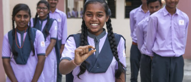 Students in purple school uniforms stand facing the camera and smiling. One girl is in front smiling with long braids and holding out a iron and folic acid supplement.