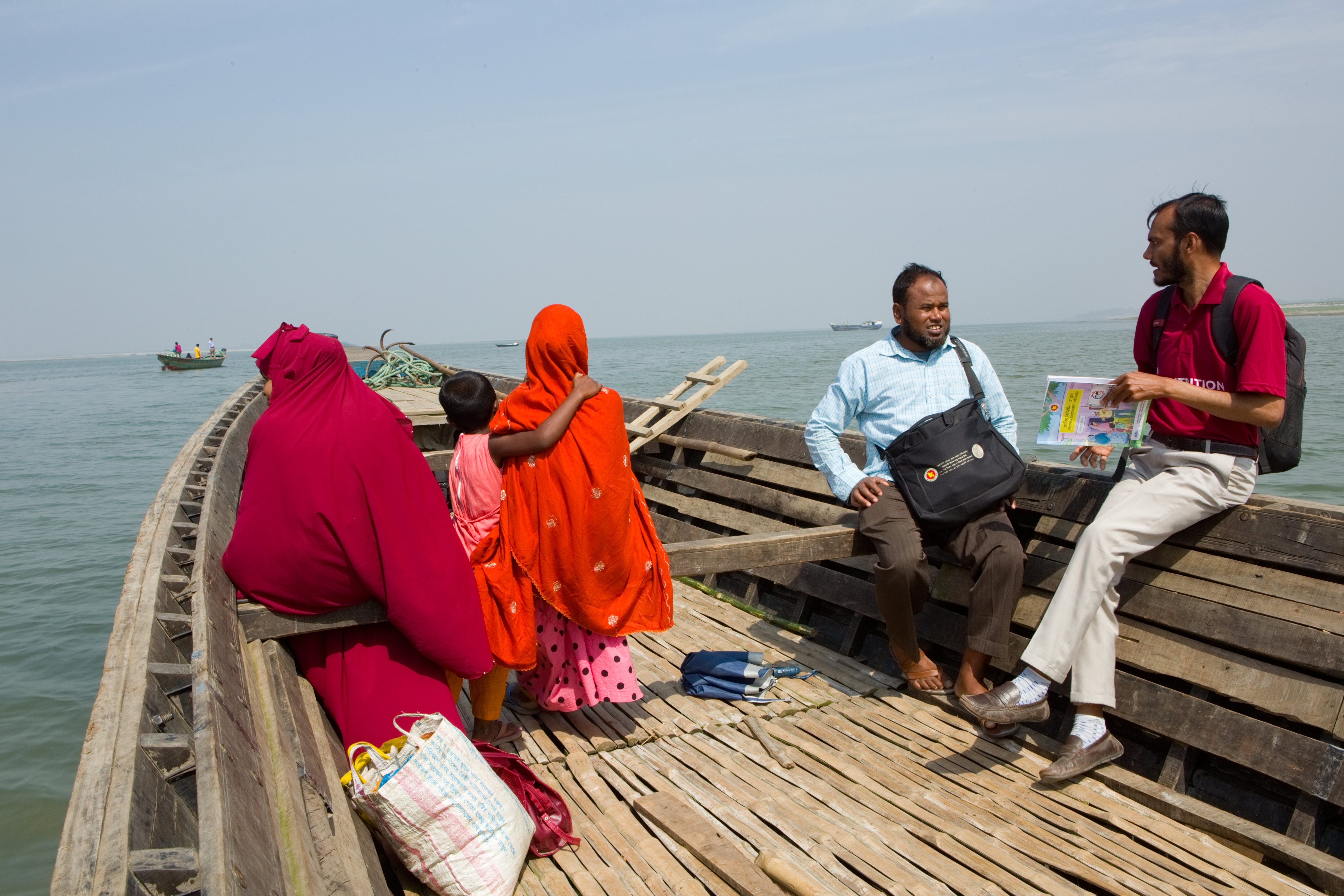 Four people sit in a wooden boat as it travels across the lake. Two people are in sarees with their backs to the camera. The other two are men in conversation and facing the camera.