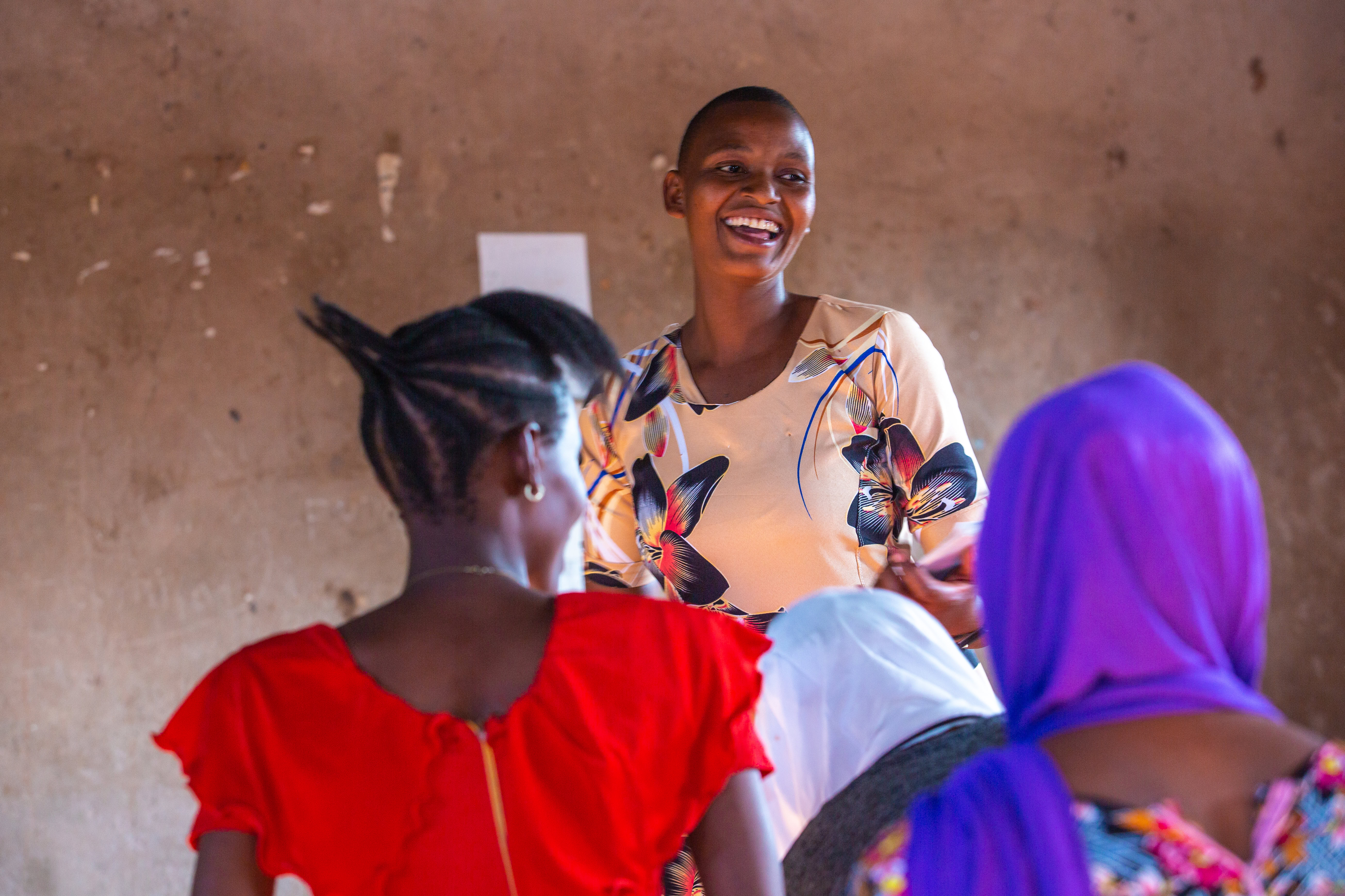 A woman addresses her peers. She is smiling while speaking to the group. We see the backs of two women listening to her.