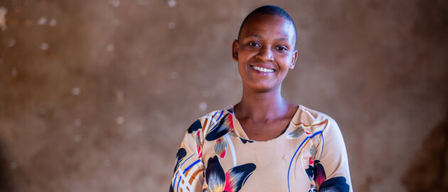 A woman stands facing the camera. She is wearing a patterened, colorful blouse and smiling to the camera.