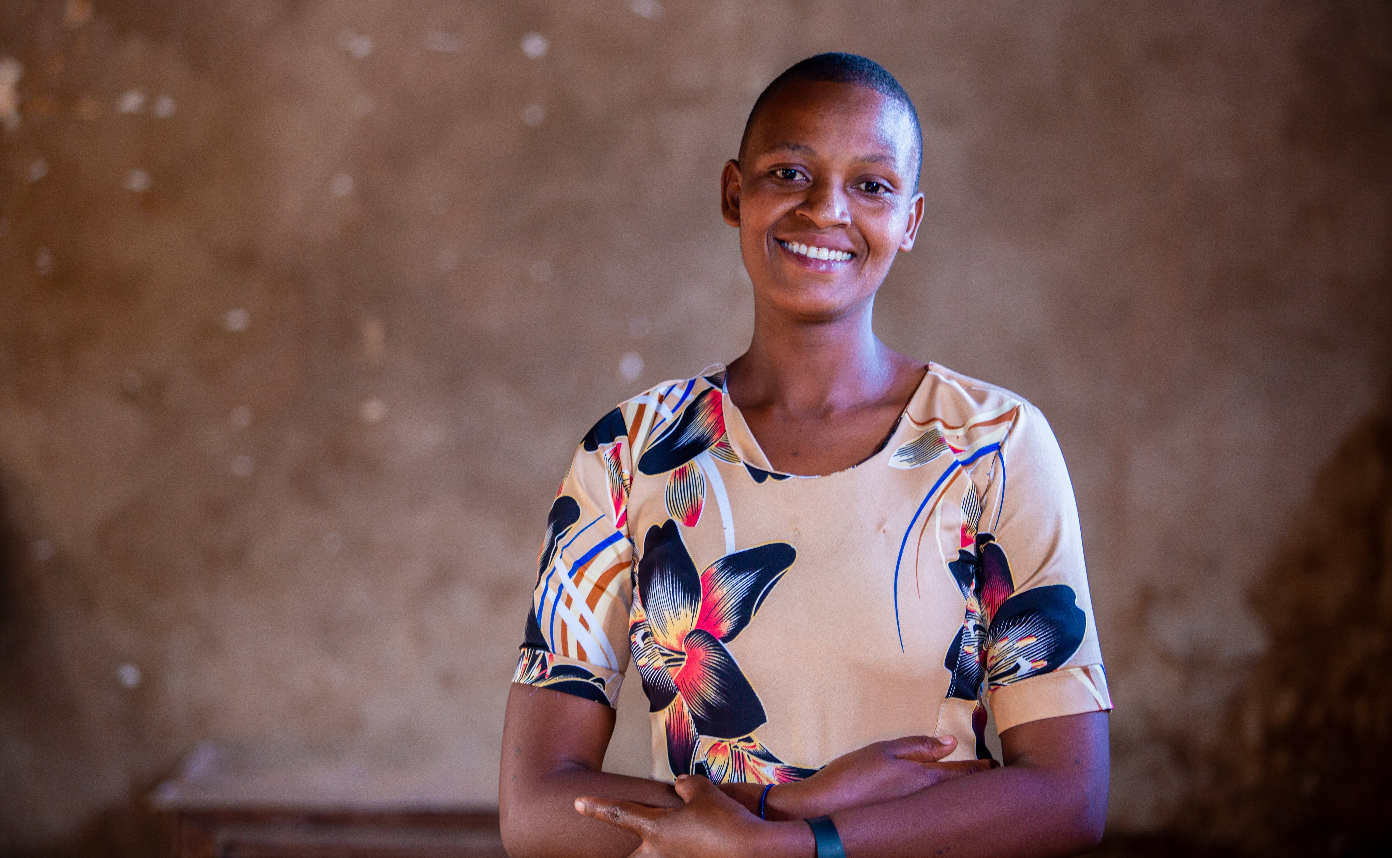 A woman stands facing the camera. She is wearing a patterened, colorful blouse and smiling to the camera