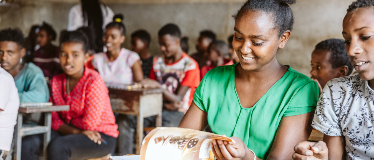 A girl laughs while flipping through a notebook. She is seated at a desk in classroom. Other students sit beside her.