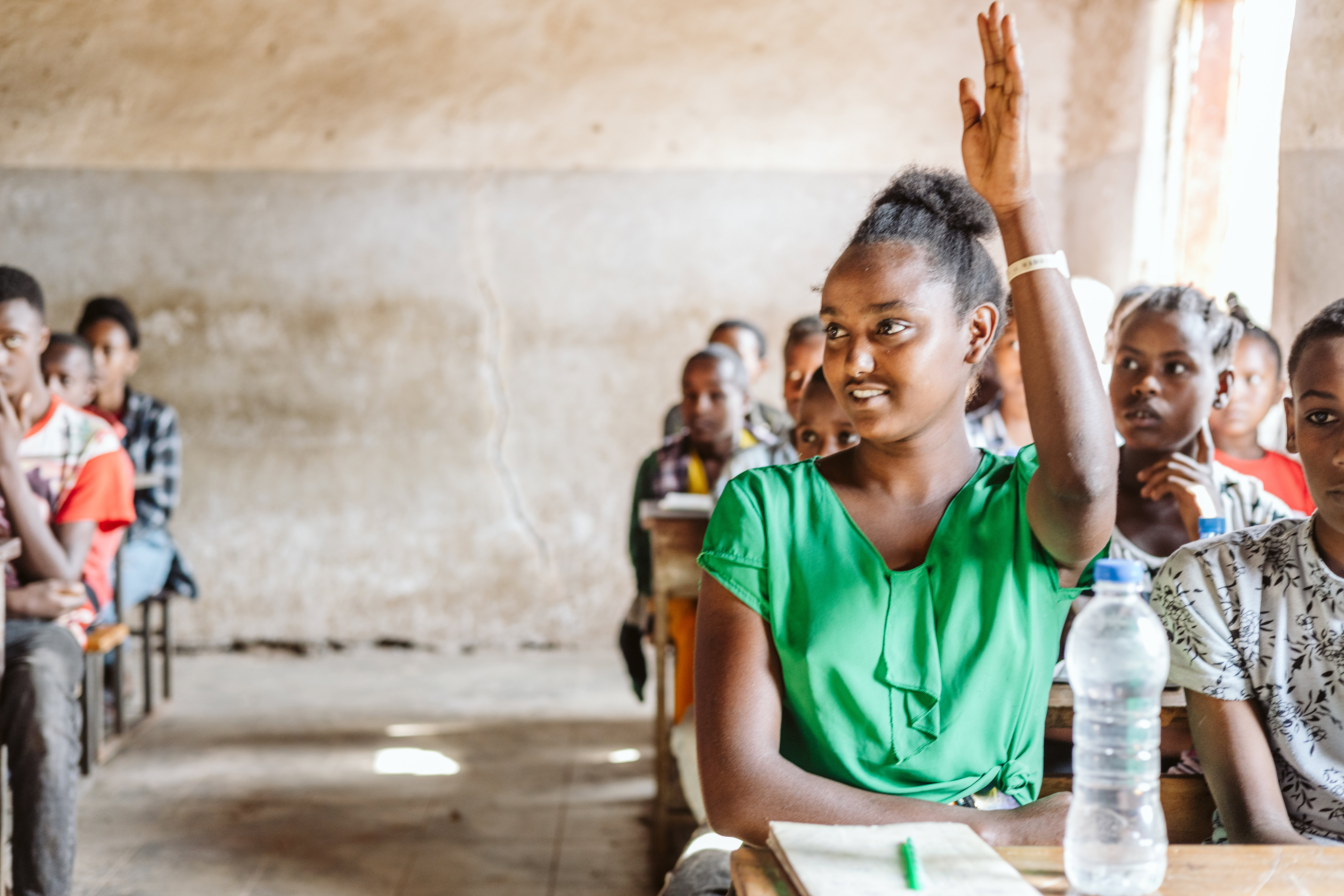 A student raises her hand. She is sitting at her desk in a classroom.