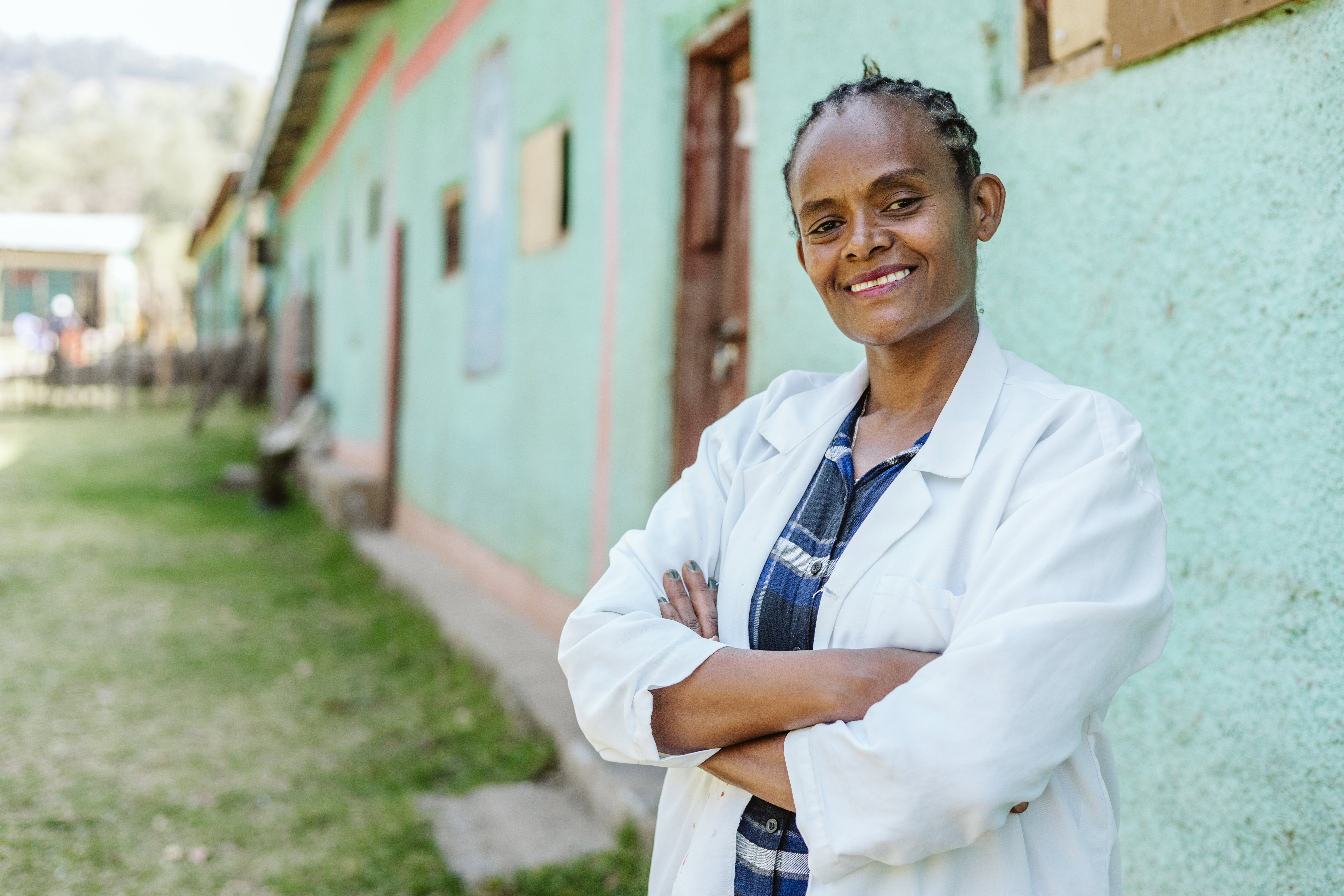 A woman in a white medical jacket stands outside. She is smiling to camera and has her arms crossed.