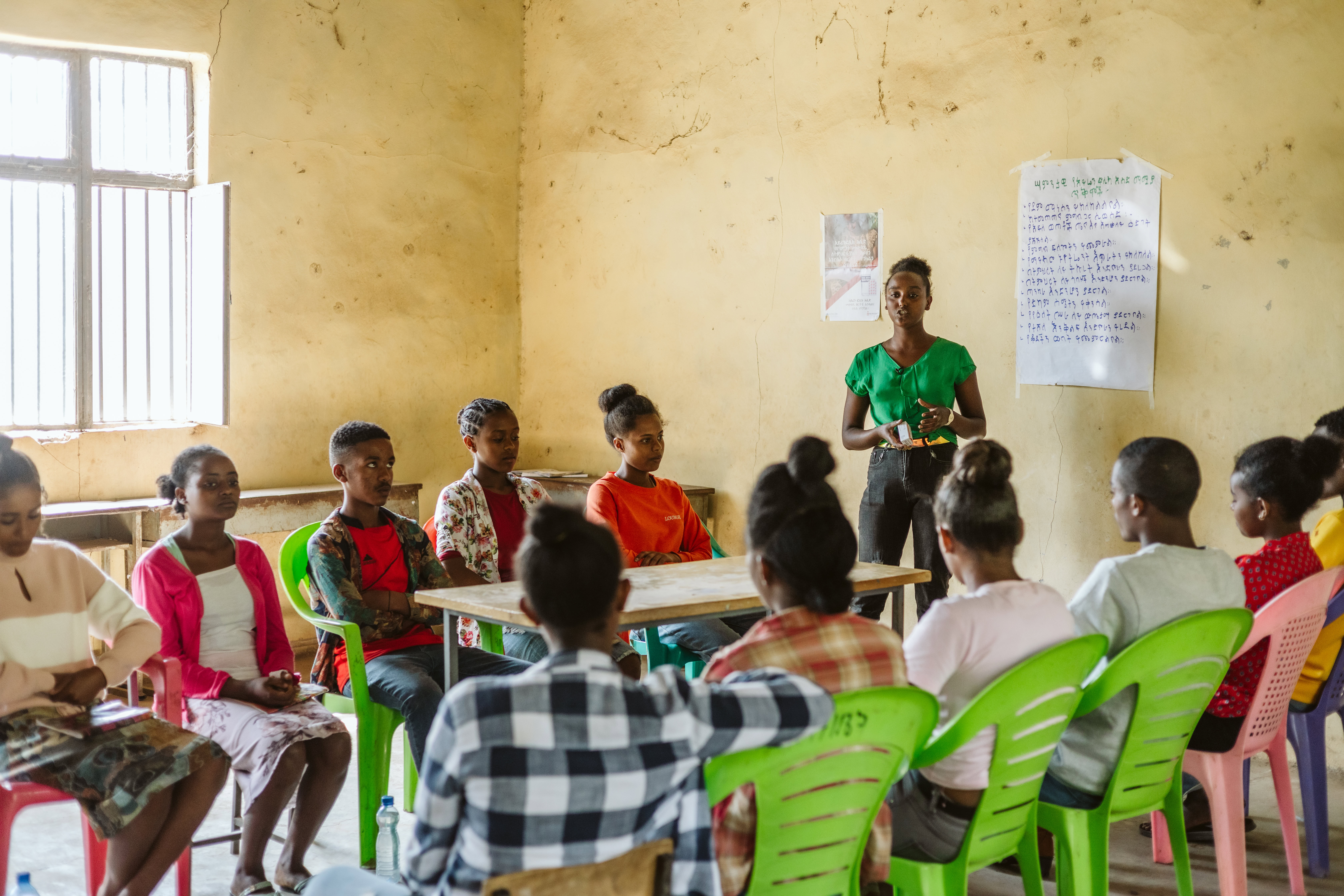 Students sit on chairs in a classroom setting in cirlce. One girl is standing addressing the group.