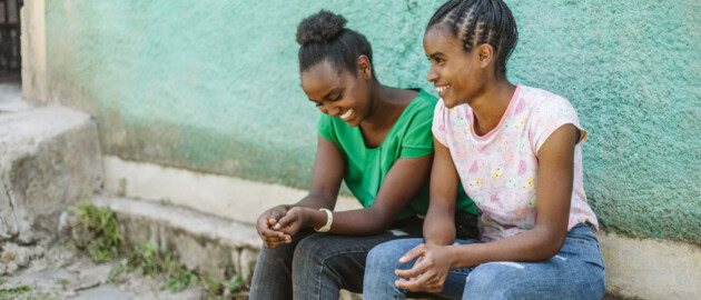 Two girl sit on bench outside. They are side by side and laughing.