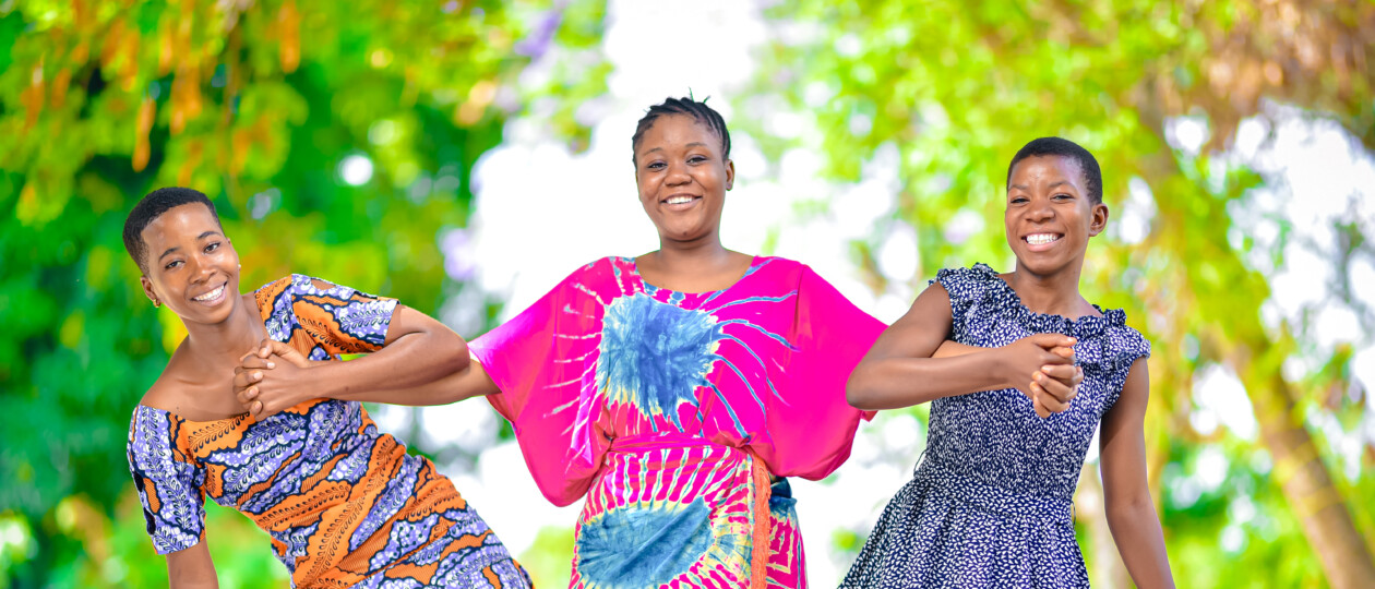 Three girls smile and laugh while standing amongst trees outside.