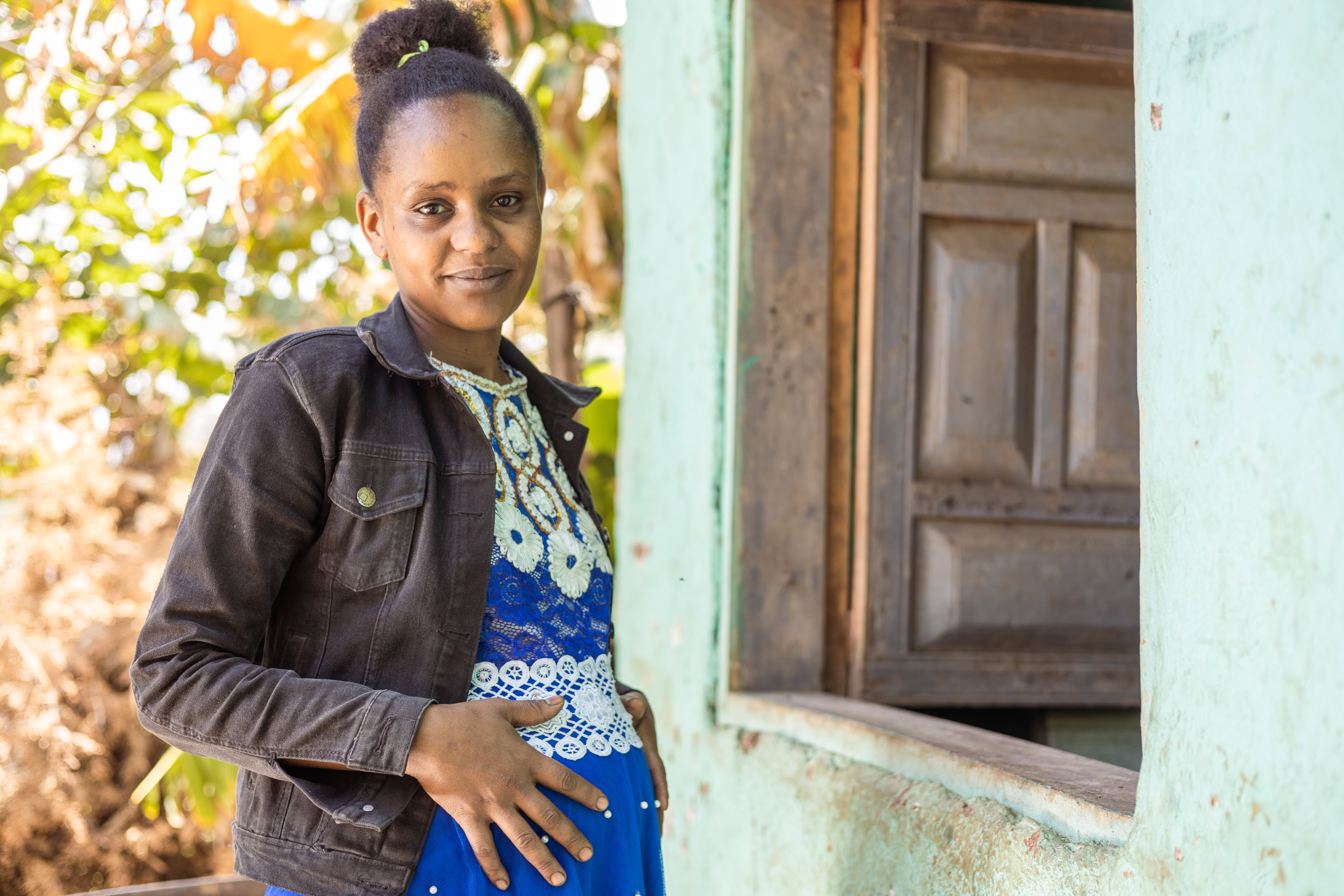 A pregnant woman stands outside. She is smiling to camera and her hands are on her stomach. 