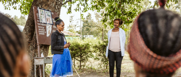 A pregnant womn is explaining her experience during a pregnant women's conference in Ethiopia. She is standing at the front of a crowd of pregnany women, seated in chairs outside.