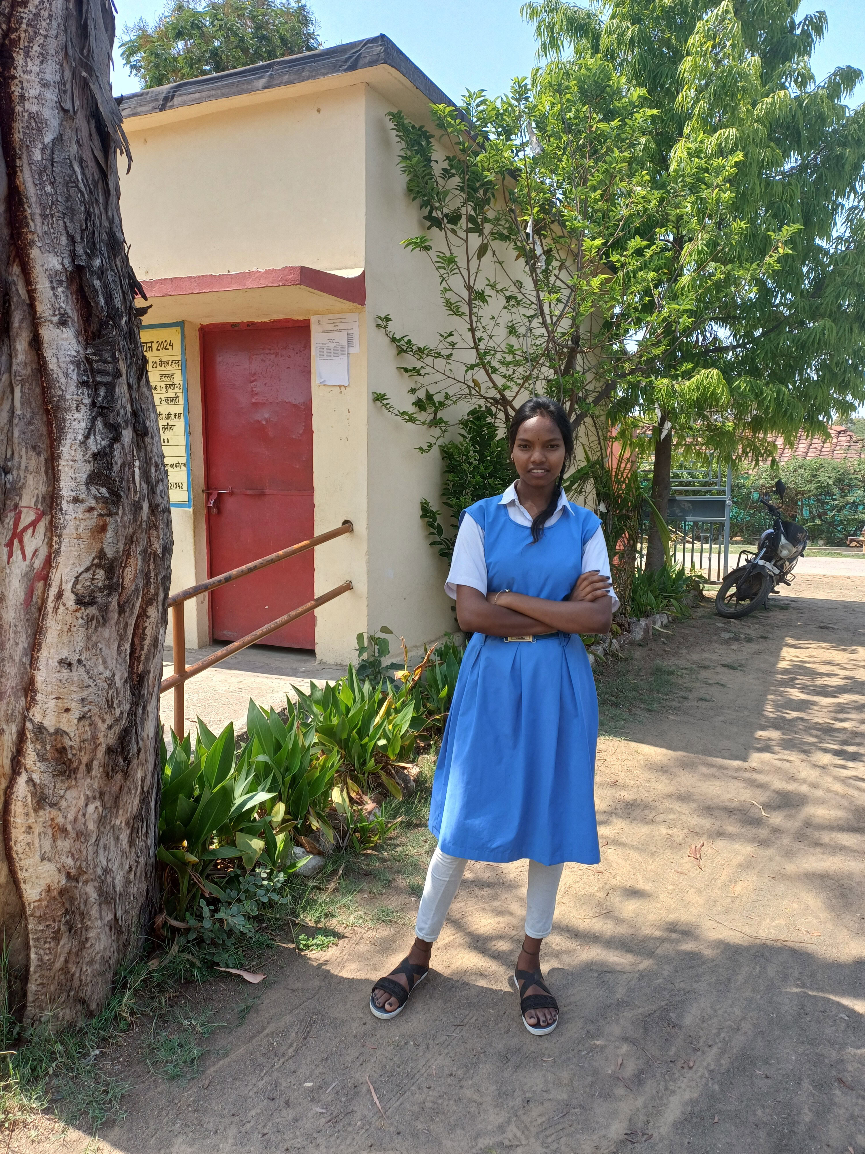 A girl stands outside in her school uniform, smiling to the camera.