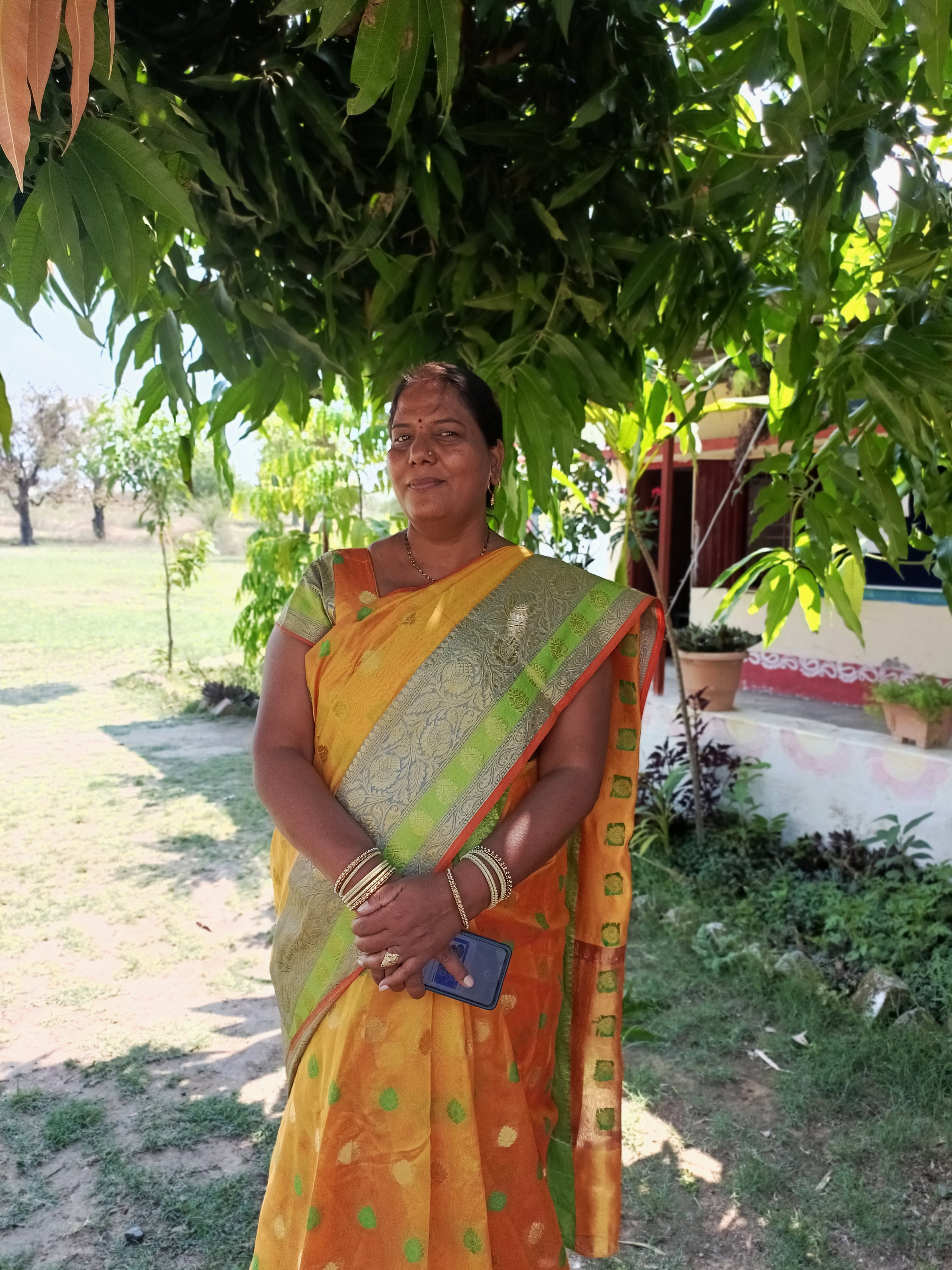 A woman stands outside looking at the camera. She is in front fo a tree in a school yard and wearing a saree.