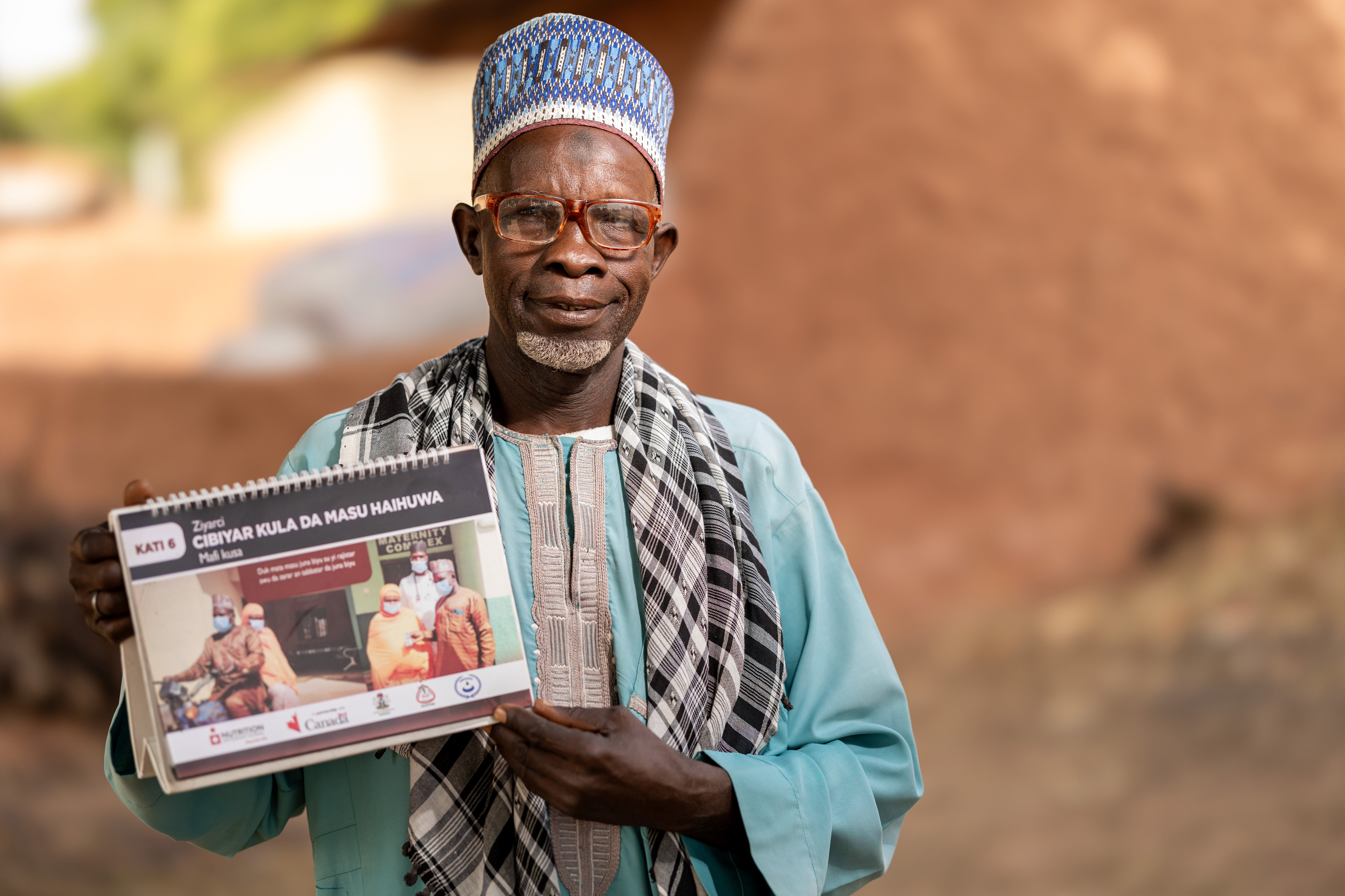 A man sits on a mat holding up a booklet on maternal health.