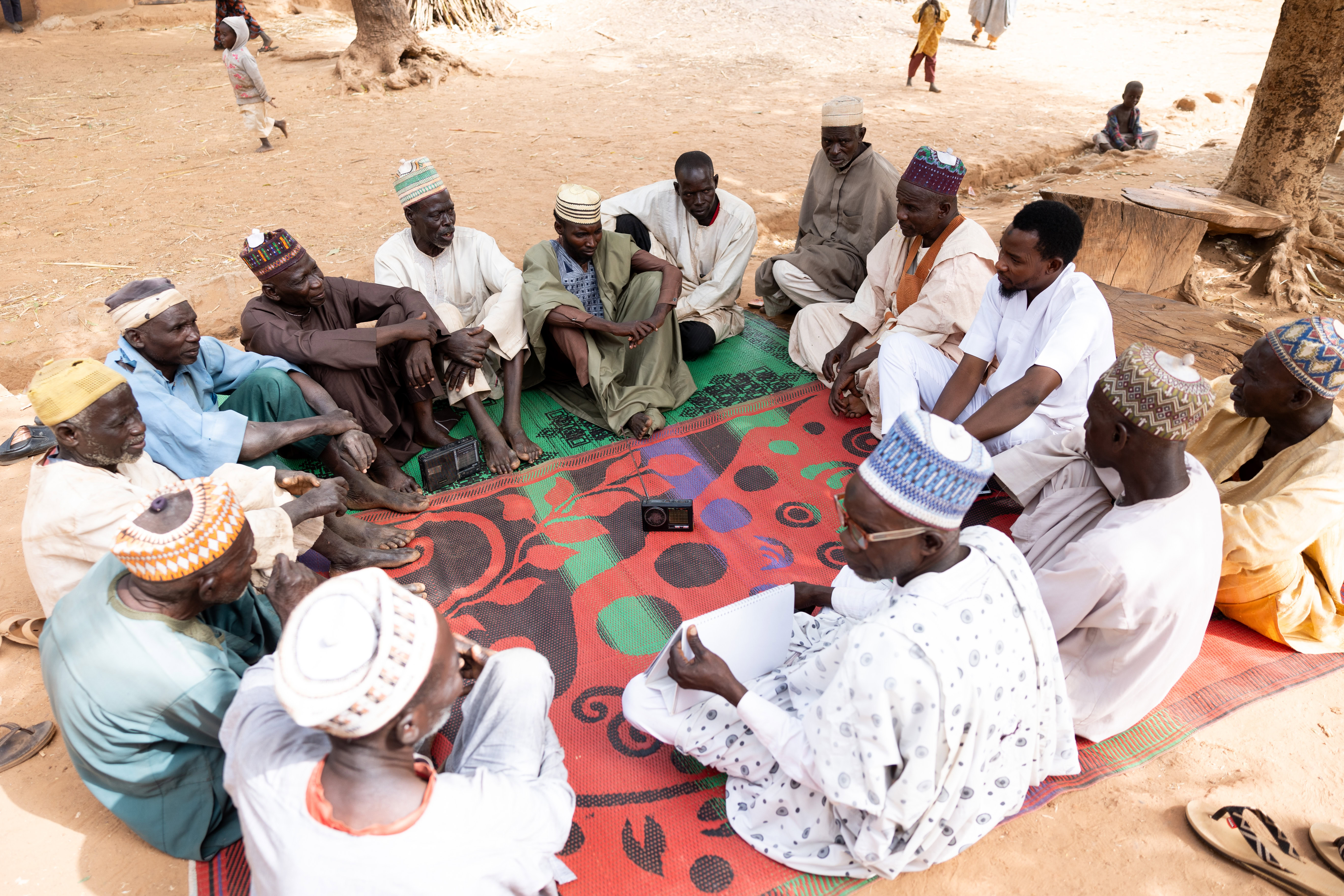 A group of men sit crossed legged on a mat outside in discussion.