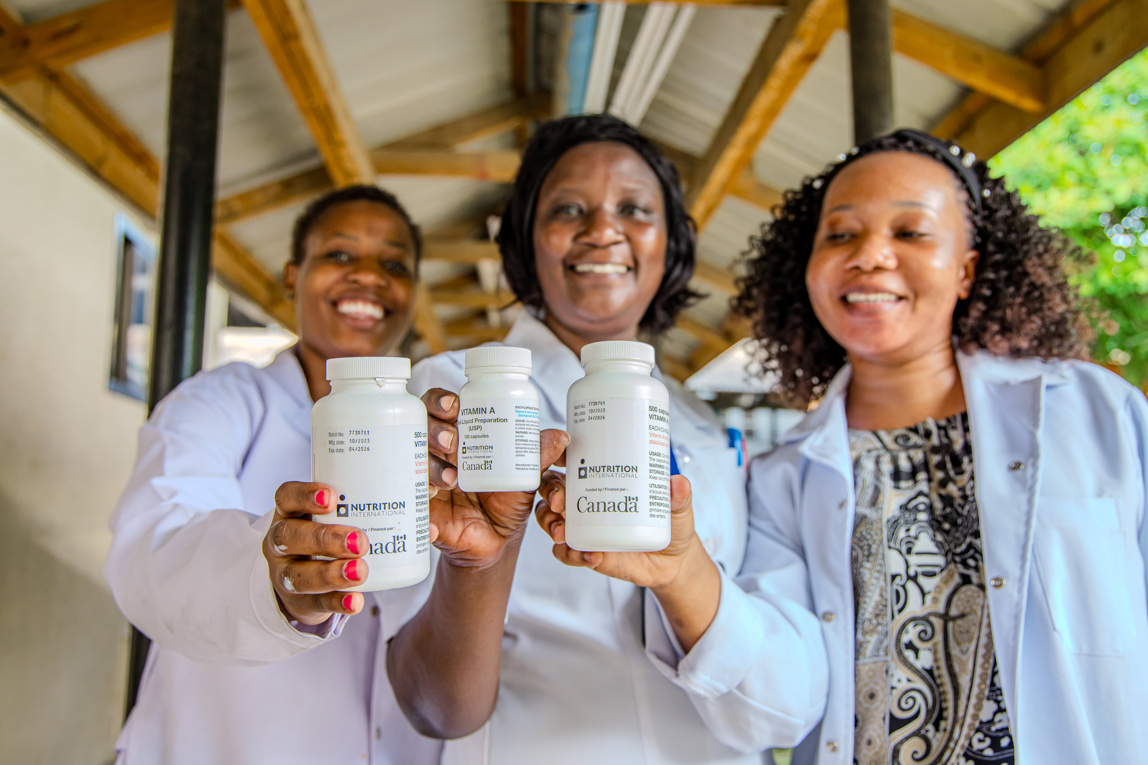 Three women stand facing the camera, each holding up a 100-count bottle of vitamin A capsules.