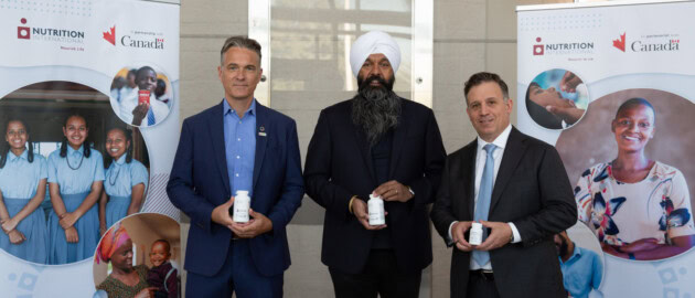 Three men stand in suits proudly holding up bottles of vitamin A supplements. They are inside. On either side are banners with logos of Nutrition International and the Government of Canada.