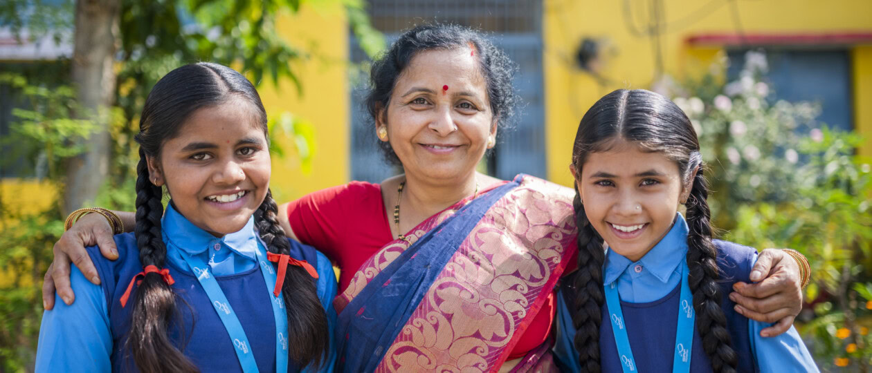 Two students stand on either side of a teacher. They are outside and smiling to the camera.