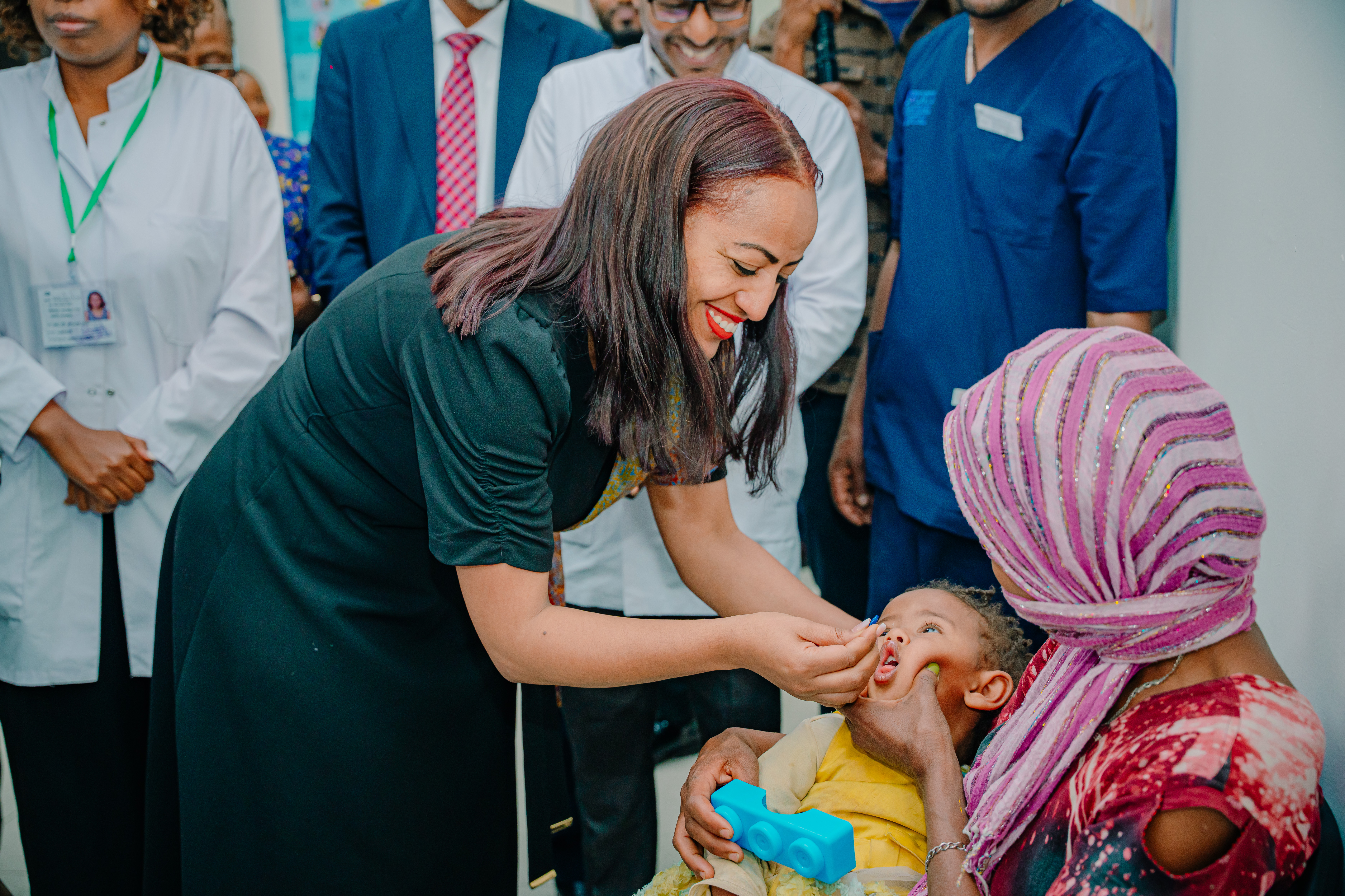 A smiling woman administers vitamin A supplementation to a child in its mother's arms.
