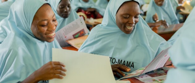 Women wearing blue mama-to-mama uniforms flip through booklets and smile, while seated in a classroom.