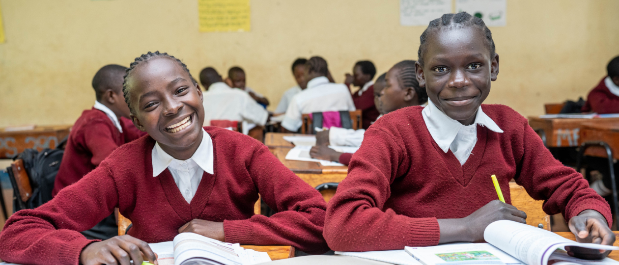 Girls sitting at a desk in a classroom wearing red school uniforms and smiling.