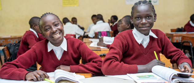 Girls sitting at a desk in a classroom wearing red school uniforms and smiling.