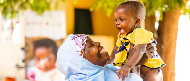 Caregiver in a blue outfit lifts a laughing toddler in a yellow shirt outdoors under trees.