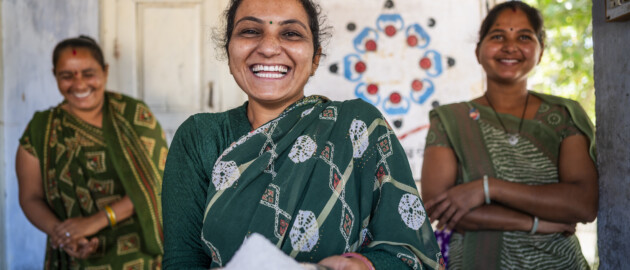 Smiling woman in a green sari holds a metal bowl with white powder, flanked by two women in patterned dresses indoors.