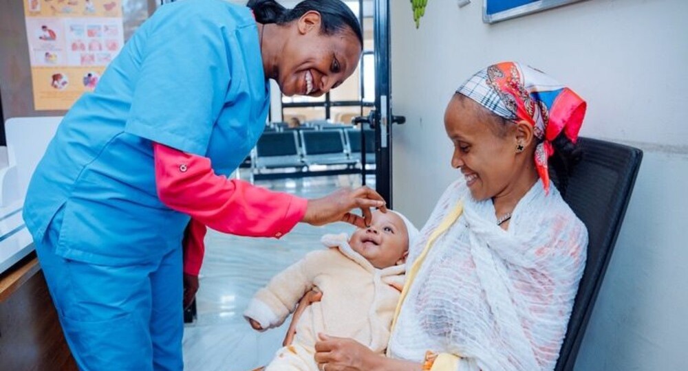 Nurse in blue scrubs gently tending a smiling baby held by a seated mother in a clinic corridor.