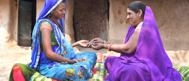 Two Indian women in blue and purple saris sit on a bright patterned blanket outside a rustic home, exchanging a blister pack of pills.